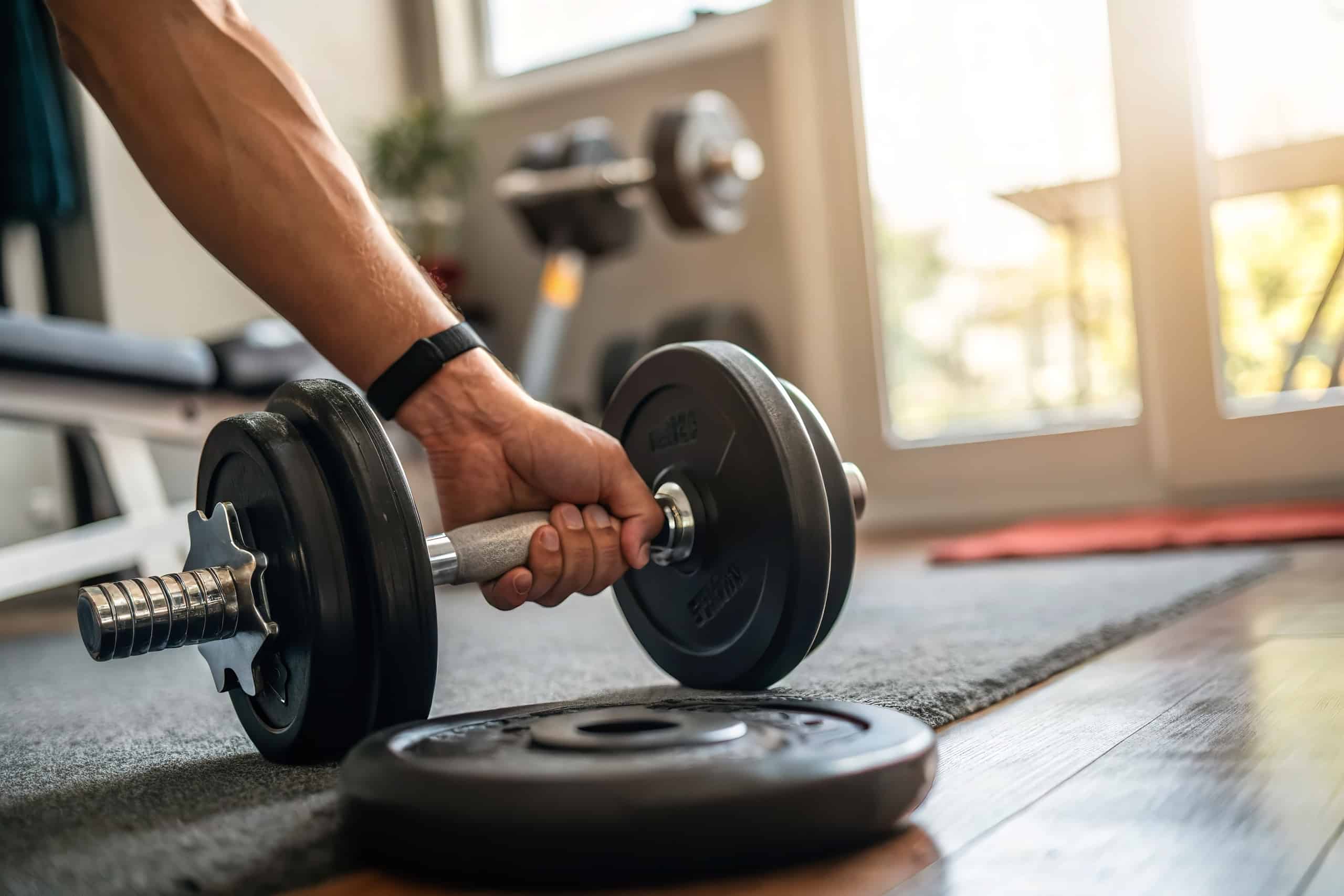 Ein Mann, der nach einer am Boden liegenden Hantel im hauseigenen Fitnessraum greift.